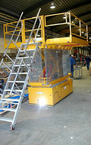 Plataforma de trabajo en posición elevada, plataformas laterales abatidas con barandillas abiertas y escalera de acceso desplegada.